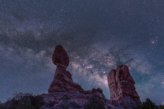 View of Balanced Rock in Arches National Park under a starry night sky with the Milky Way galaxy visible above the red sandstone formations in Moab, Utah, United States.