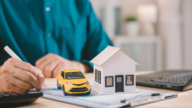 Person shows financial planning, responsibility, income management for household expenses with house model and yellow car, using calculator while writing on report at wooden office desk