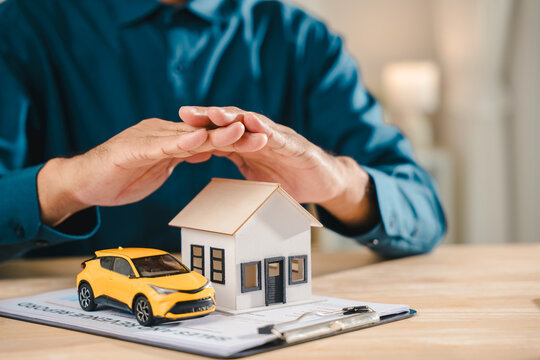 Insurance concept life home auto protection shown through male hands shielding house model and small yellow car on office desk, providing financial security and property insurance for family