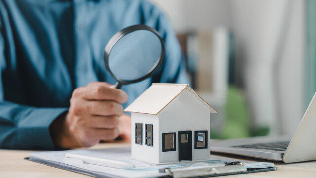 Professional agent holds magnifying, house model sits on office desk, expert inspector checking property details for home mortgage, buyer searching residential building for future investment