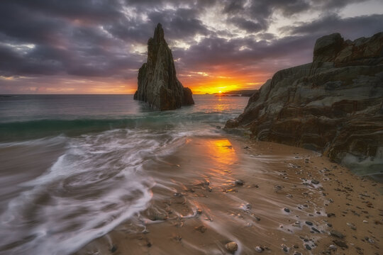 View of a dramatic sea stack rising from the ocean at sunset with waves washing over the sandy beach in Asturias, Spain.