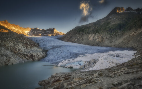 View of the Rhone Glacier with its icy surface, a turquoise meltwater lake, and surrounding rocky mountain peaks under golden hour light in Switzerland.