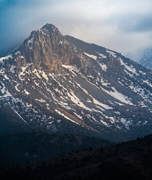 View of the snow-capped peaks of the Zaamin Mountains, featuring rugged rocky slopes, patches of snow, and dramatic clouds at sunset in Zaamin, Jizzakh Region, Uzbekistan.