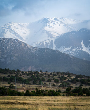 View of the snow-capped peaks of the Zaamin mountains rising above the juniper-dotted foothills under a cloudy sky in Zaamin, Jizzakh Region, Uzbekistan.