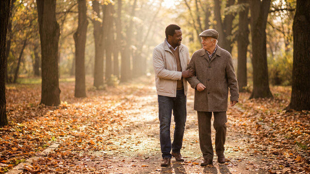 Adult son holding elderly father arm while walking down sunny park autumn pathway filial piety photo