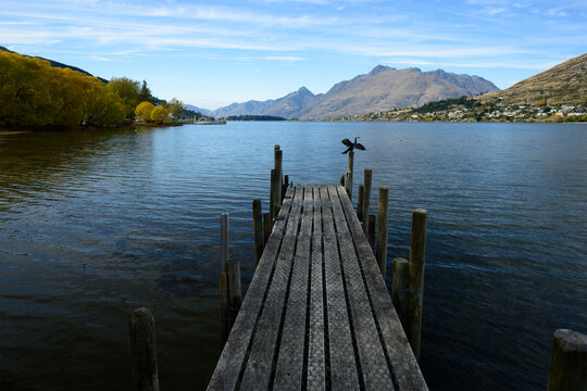 Wooden Pier on Tranquil Lake: A weathered wooden pier stretches across a calm, reflective lake, leading towards distant, majestic mountains under a clear blue sky. A lone bird perches on a post.