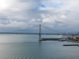 Obraz premium San Francisco Bay Bridge cables crossing the bay waters with the city skyline and cloud-filled sky providing an iconic California cityscape view from above
