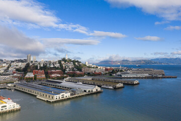 Obraz premium Panoramic aerial view capturing San Francisco's historic Embarcadero waterfront with piers, city architecture, Coit Tower, and the Golden Gate Bridge in the distance under a blue sky