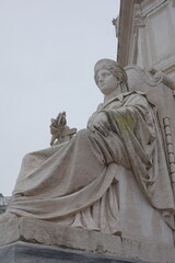 Fototapeta premium Low angle close-up of classical robed stone female figure sculpture on historic building facade holding cross against gray overcast sky in Lisbon