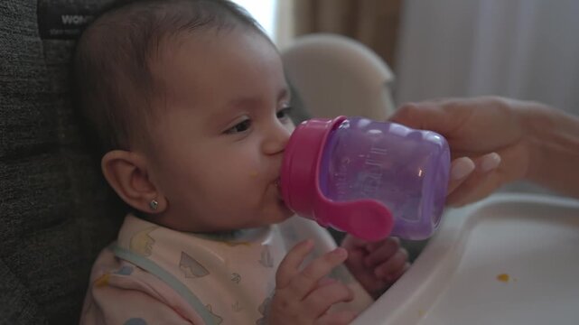 Parent helps a baby drink water from a purple sippy cup while sitting in a high chair, ideal for hydration, infant care, feeding routines and parenting lifestyle themes.