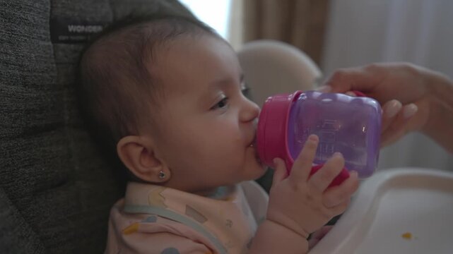 Parent helps a baby drink water from a purple sippy cup while sitting in a high chair, ideal for infant care, hydration, feeding routines and parenting lifestyle themes.