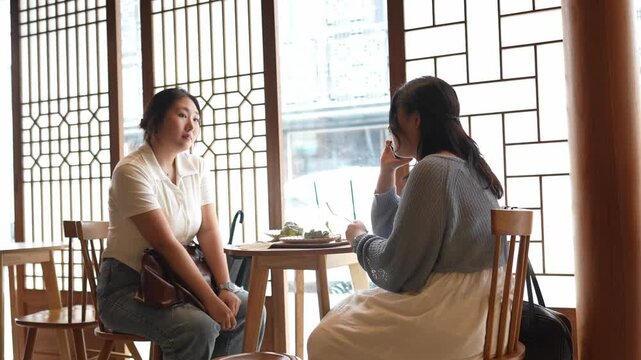 Two women converse softly at a wooden table. Their calm expressions suggest deep, thoughtful conversation. Natural light streams through traditional lattice windows