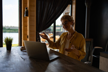 Naklejka premium A Caucasian woman with short blonde hair and glasses is actively engaged in a video call on her laptop, gesturing with her hands while sitting at a wooden table