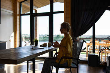 Naklejka premium A Caucasian woman sits at a wooden table, engaged in a video call on her laptop with a scenic view of a lake and greenery in the background