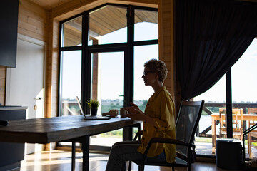 Naklejka premium A Caucasian woman wearing glasses and a yellow shirt is sitting at a wooden table in a modern log cabin, engaged in a video call on her laptop
