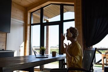 Naklejka premium A Caucasian woman wearing headphones and glasses is engaged in a video call on her laptop while sitting at a wooden table in a room with large windows