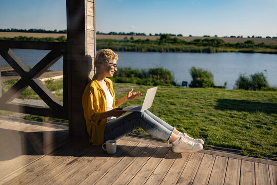 A Caucasian woman with short blonde hair wearing glasses and a yellow shirt is sitting on a wooden porch with a laptop and a mug of coffee