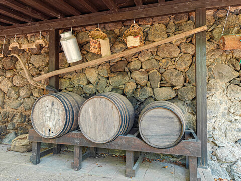 Wooden barrels are positioned on a sturdy wooden frame against a rustic stone wall. Above them, a long wooden beam extends horizontally, supporting various items. 