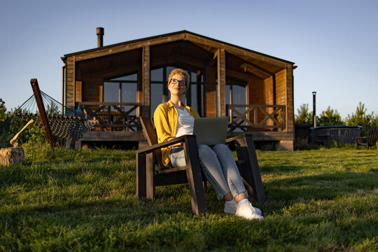 A young woman with short blonde hair and glasses sits in a chair outdoors with a laptop on her lap, enjoying the sunshine in front of a wooden cabin