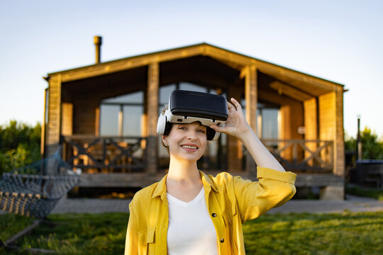 A smiling caucasian woman wearing a yellow shirt and VR headset stands in front of a wooden house with a hammock nearby