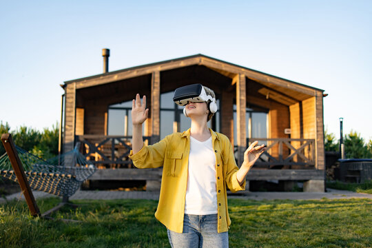 A young woman wearing a virtual reality headset and headphones enjoys an immersive experience in a natural outdoor setting near a wooden cabin