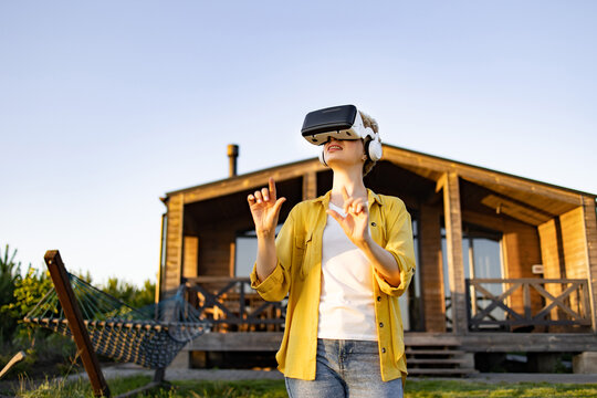 A young caucasian woman wearing a virtual reality headset and headphones enjoys an immersive experience in a natural outdoor setting near a wooden house