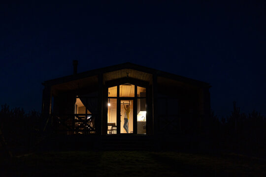 A young caucasian woman dances joyfully inside a contemporary wooden cabin at night, illuminated by warm interior lights