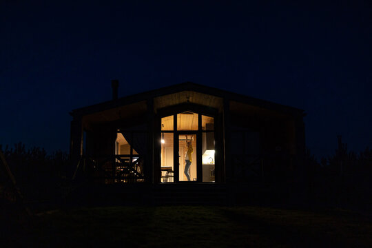 A lone caucasian woman dances joyfully inside a modern wooden cabin at night, with warm interior lights creating a cozy ambiance