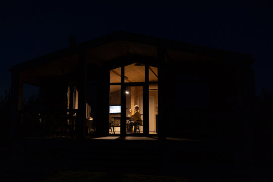 A solitary caucasian woman works on her laptop inside a cozy wooden cabin illuminated by warm interior lights against the dark night sky