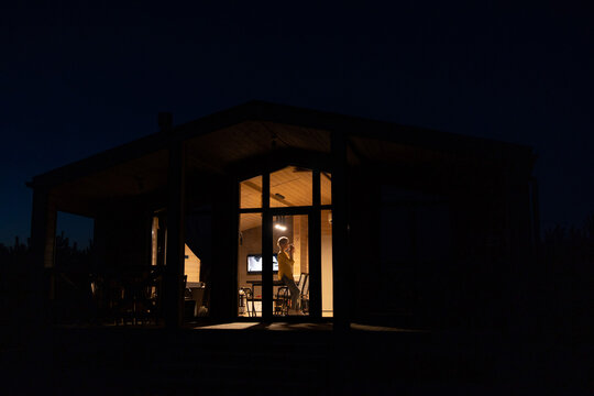 A solitary woman stands inside a warmly lit cabin at night, enjoying a quiet moment