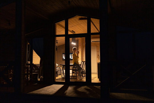 A caucasian woman stands inside a warmly lit cabin, holding a mug and looking out into the dark night