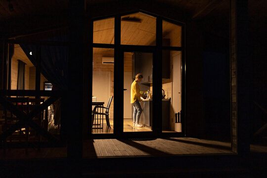 A caucasian woman stands in a modern kitchen preparing food late at night, illuminated by interior lights