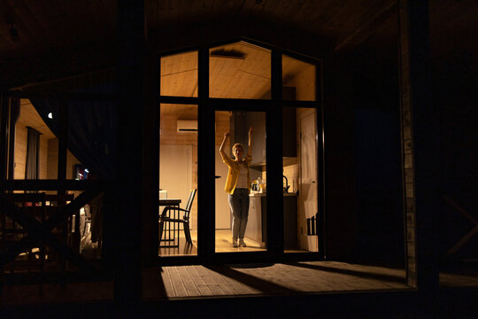 A young caucasian woman joyfully dances inside a brightly lit modern cabin kitchen at night, seen through large glass doors