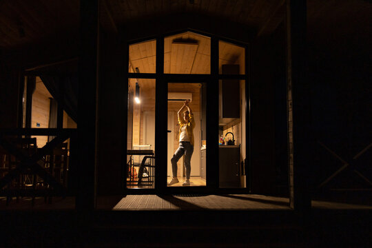 A young caucasian woman dances joyfully inside a modern wooden cabin at night, illuminated by warm interior lights