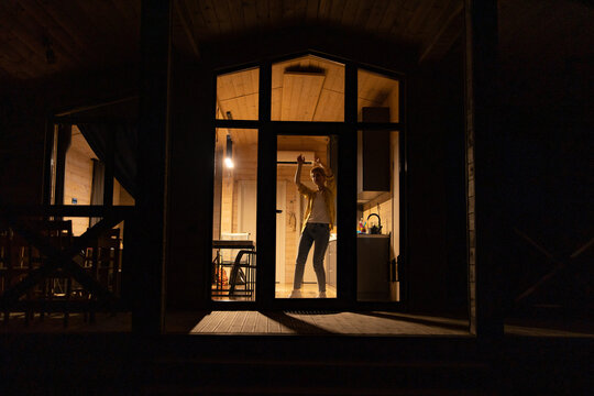 A young caucasian woman joyfully dances inside a modern wooden cabin kitchen at night, illuminated by warm interior lights
