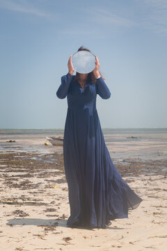 woman in long blue dress on the beach hiding behind round mirror with reflection of the sky