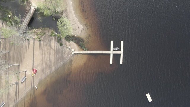 Aerial view of Commanda Lake showing a wooden dock extending into the dark water with a small boat moored alongside, sandy shoreline, and a bridge in Restoule, Ontario, Canada.