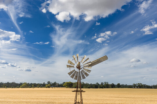 Aerial view of puffy white clouds in a blue sky above a farm windmill in the middle of a dry paddock