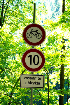 Slovak traffic signs on a metal pole forbidding cycling, setting a 10 km/h speed limit, against green summer trees.