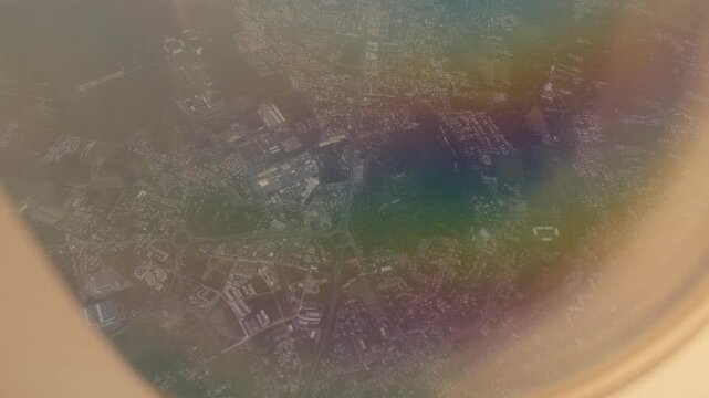 Aerial footage of an urban settlement taken through an airplane window during flight. The scene shows industrial and residential areas with hazy atmospheric conditions, captured from high altitude wit