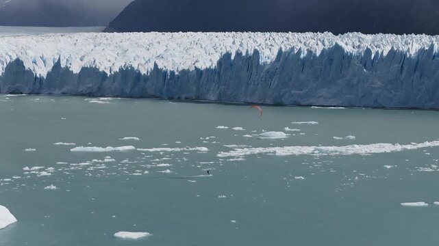 Kitesurfer on Lago Argentino near Perito Moreno Glacier, Los Glaciares National Park, Santa Cruz, Argentina, moves across cold waters beneath a towering ice wall and floating icebergs telephoto aerial