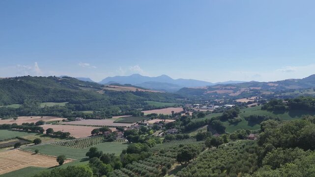 Ampio paesaggio rurale con colline verdi, campi coltivati e un piccolo centro abitato nella valle, con montagne sullo sfondo sotto un cielo azzurro limpido