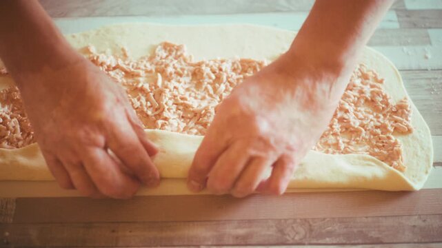 Close-up of chef hands rolling a rectangular dough sheet filled with ham and cheese into a big roll. Process of making homemade pizza pinwheels or savory swirls on wooden table background, 4k.