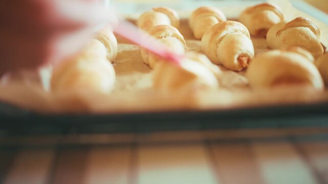 Close-up of chef hand using a pastry brush to glaze raw crescent rolls with milk on a baking tray. Preparing homemade savory pastry with ham and cheese before oven baking sequence, 4k.