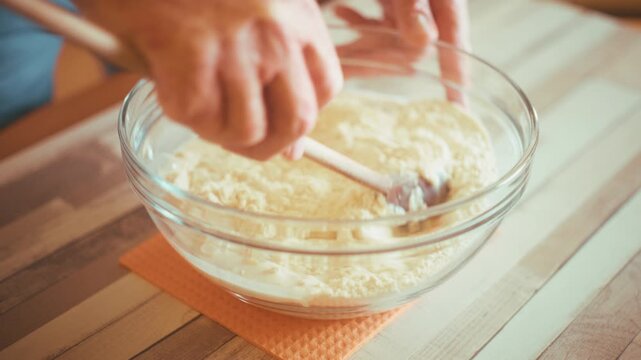 Close-up of chef hands mixing flour and ingredients with a wooden spoon in a glass bowl to make fresh dough. Preparing homemade pastry or bread on a wooden kitchen table background, 4k.