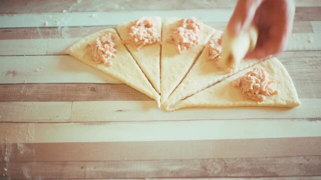 Close-up of chef hands rolling filled dough triangles into traditional crescent rolls or croissants. Preparing homemade savory pastry with ham and cheese on a wooden kitchen table, 4k.