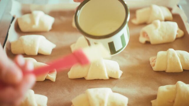 Close-up of chef hand using a pastry brush to glaze raw crescent rolls with milk on a baking tray. Preparing homemade savory pastry with ham and cheese before oven baking sequence, 4k.