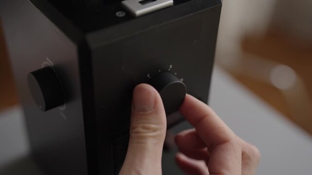 Close-up view of person hand turning adjustment knob on black electric burr grinder, precisely setting desired coarseness for brewing aromatic coffee. Shooting in slow motion