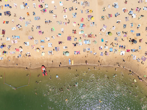 beachgoers sunbathing and swimming in tan water with umbrellas and towels under blue skies, aerial view of relaxed and joyful families enjoying summer vacation.