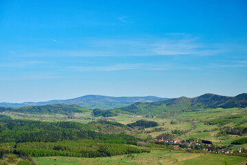 Obraz premium Wide view of green mountain valley with forests, hills and small village areas under clear blue sky. Concept of nature travel, hiking and rural landscape.